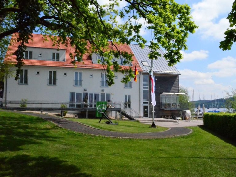 Ein modernes Gebäude mit roten Dächern, umgeben von Rasen und Bäumen, am Ufer eines Sees gelegen.